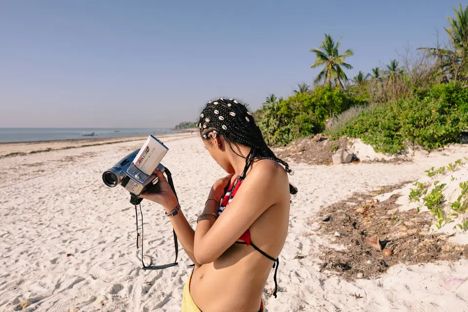 A woman with braided hair holding a vintage camcorder on a sunny tropical beach